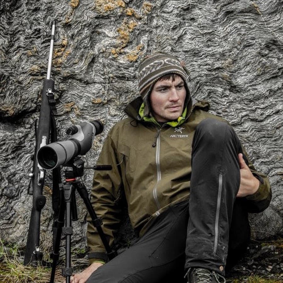 Logan Evans sitting against folded rock with a spotting scope.